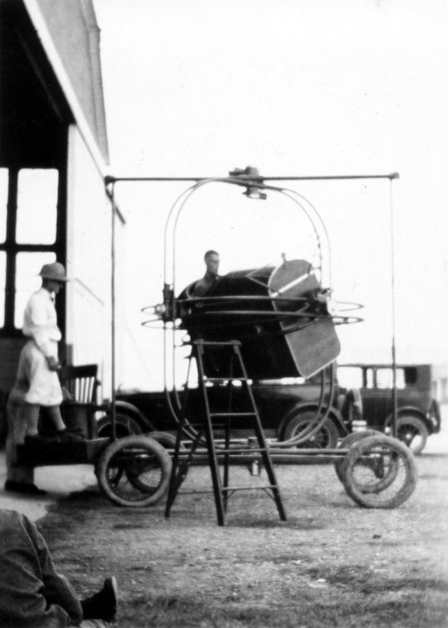 A cadet strapped into a rudimentary, mechanical multi-axis flight simulator mounted on a rolling frame.