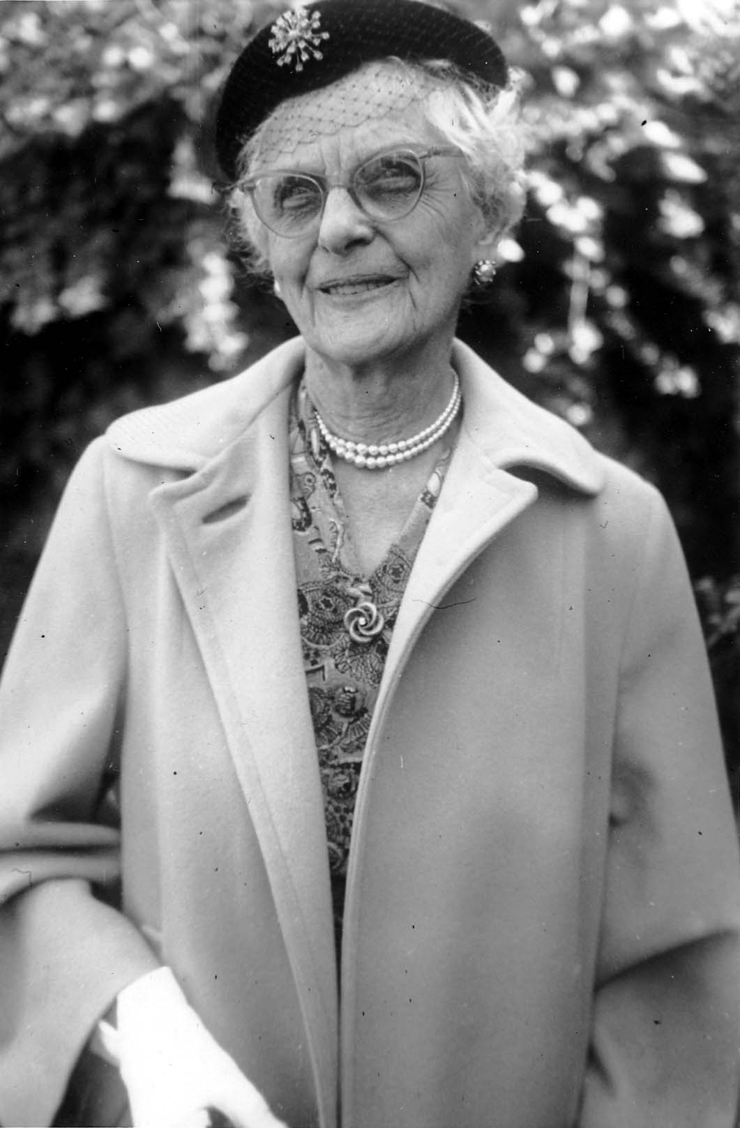 A close-up portrait of an elegant elderly woman wearing winged cat-eye glasses, a pearl necklace, and a dark pillbox hat with veil netting and a brooch.