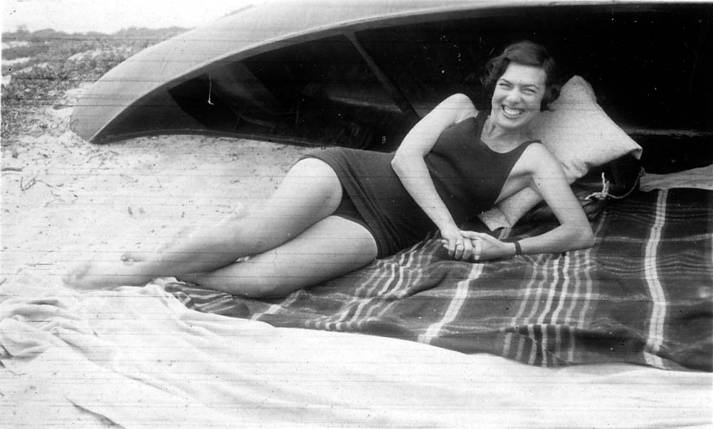A playful portrait of Elisabeth "Betty" Hall smiling broadly, wearing a dark swimsuit and reclining on a blanket in the sand next to an overturned boat.