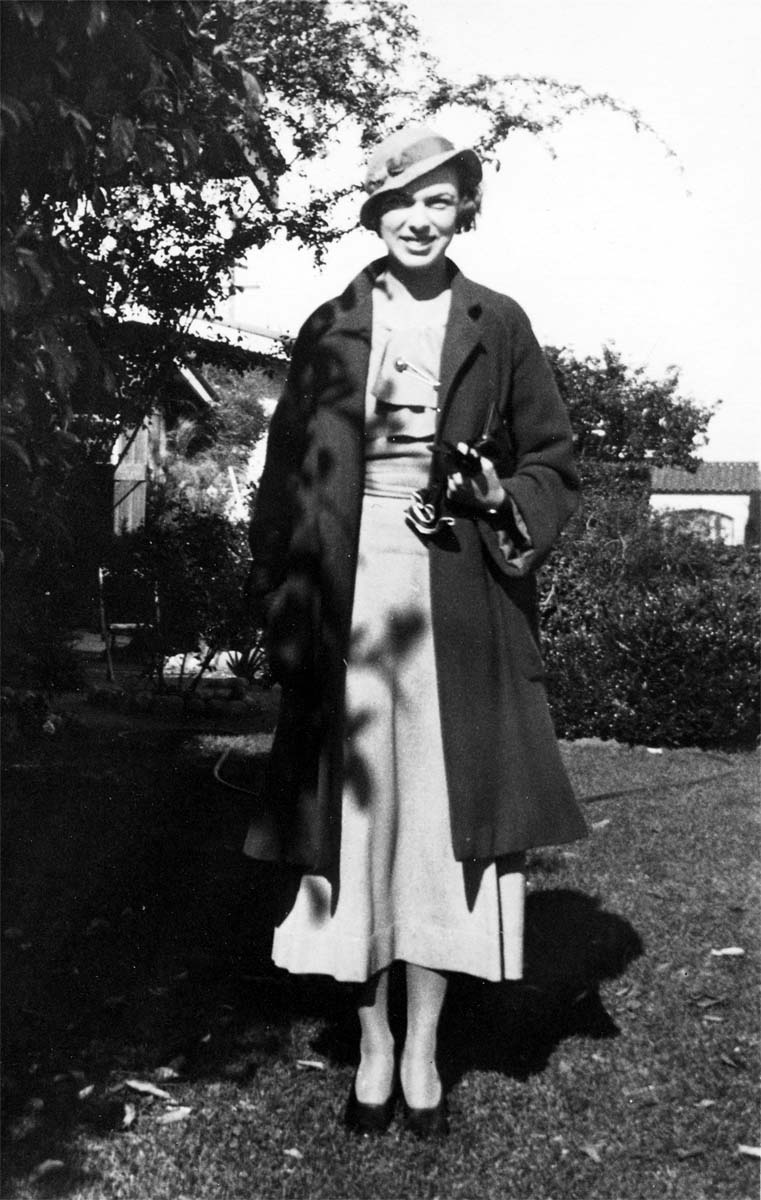A portrait of Elisabeth "Betty" Hall standing in a sunny garden, wearing a light-colored dress, a dark coat, and a wide-brimmed hat.