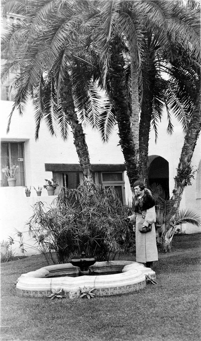 A portrait of Elisabeth "Betty" Hall standing next to an ornate stone fountain and tall palm trees in the courtyard of the Hotel Mar Monte.