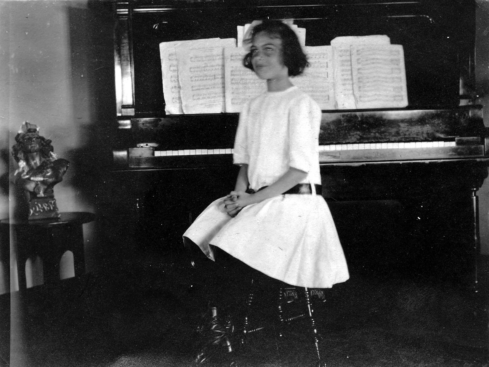 A young Elisabeth Betty Hall in a white dress seated at an upright piano with open sheet music and a sculptural bust on a nearby side table.
