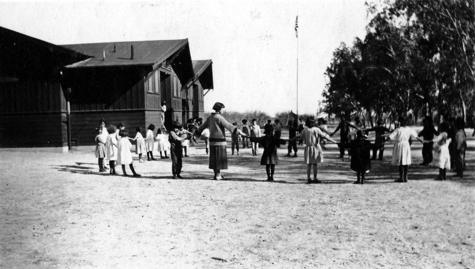 Schoolteacher Elisabeth Walker leading dozens of young schoolchildren holding hands in a large circle outside a wooden schoolhouse.