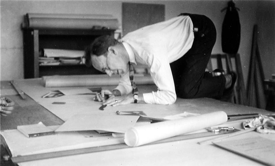 An engineer wearing a shirt and tie kneeling directly on top of a massive architectural blueprint spread across a large drafting table.