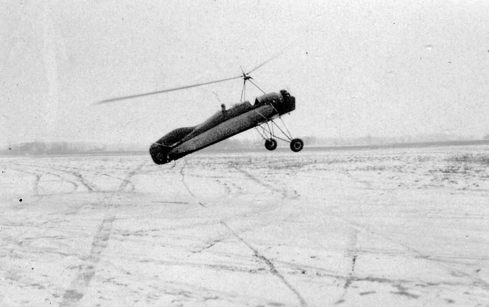 An experimental autogyro flying low over a snow-covered field marked with tire tracks.