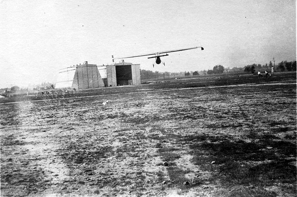 A very lightweight, high-wing experimental aircraft or glider flying low over a grassy airfield, with large hangars in the background.