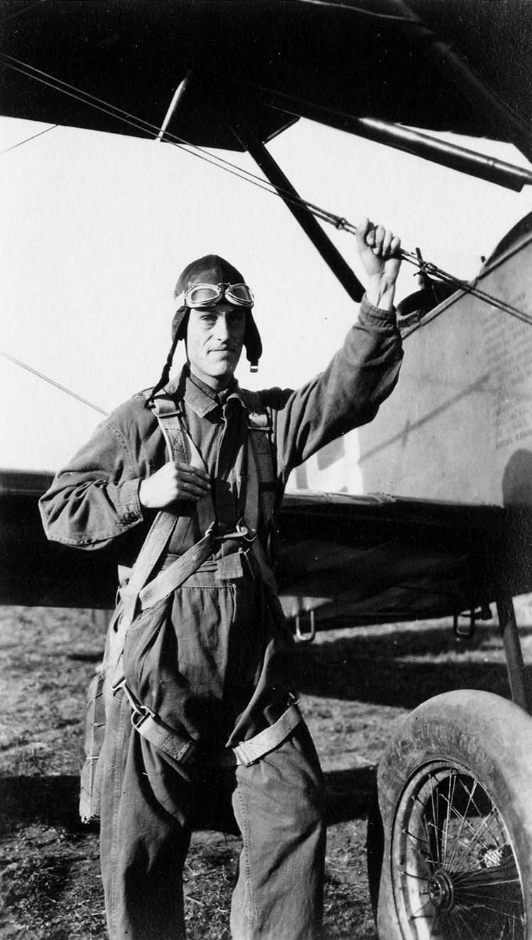 An Army Air Corps flight cadet in a flight suit and leather helmet, holding a bracing wire next to a training biplane.