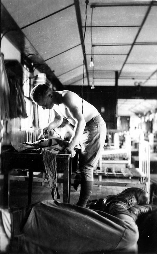 A shirtless flight cadet standing in the barracks, using a heavy iron to press his uniform pants on a small wooden table while another cadet sleeps nearby.