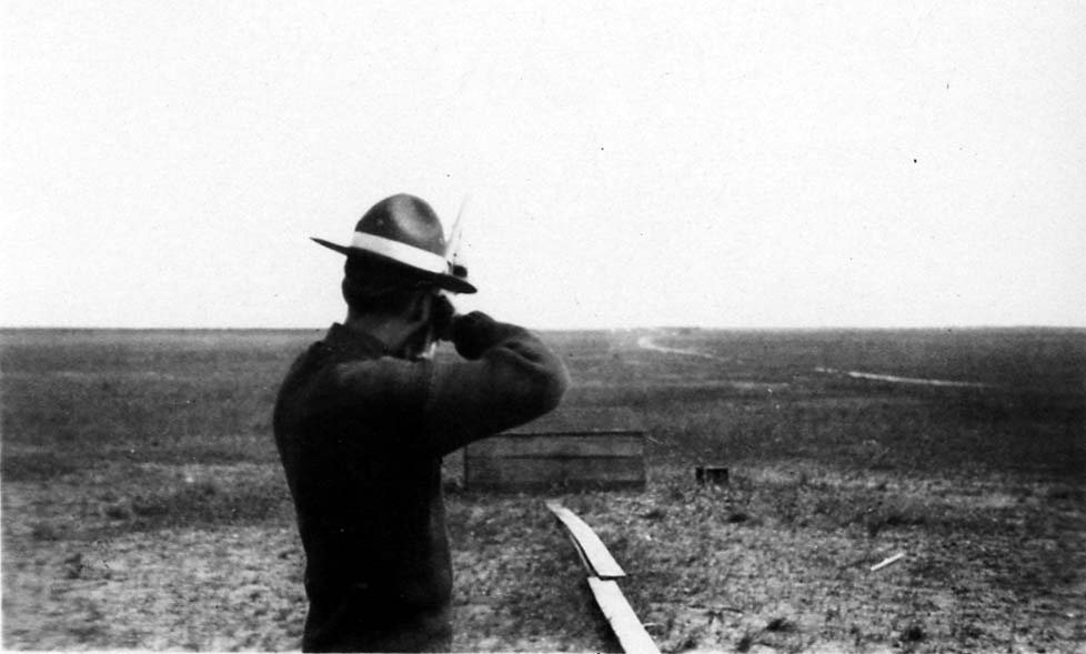 A flight cadet wearing a campaign hat and coveralls, seen from behind, looking through a sighting instrument across a vast, flat landscape.