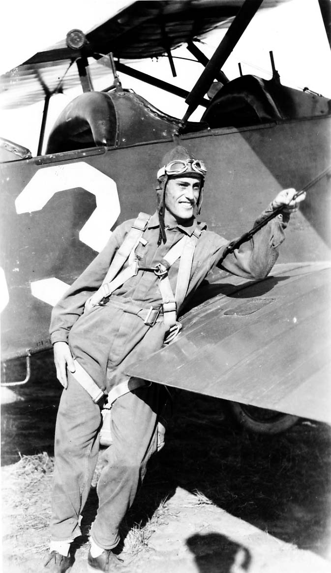 An Army Air Corps flight cadet posing casually against the lower wing of a training biplane, wearing his leather flying cap and goggles.