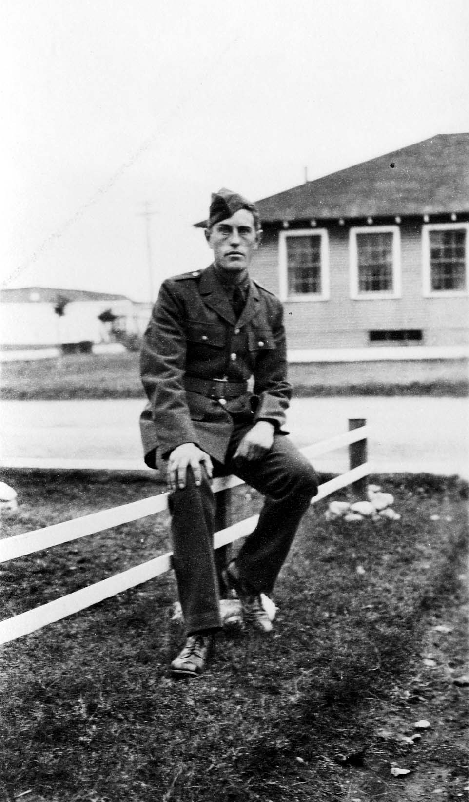 A young Army Air Corps flight cadet in a crisp dress uniform and overseas cap sitting casually on a low white wooden fence.