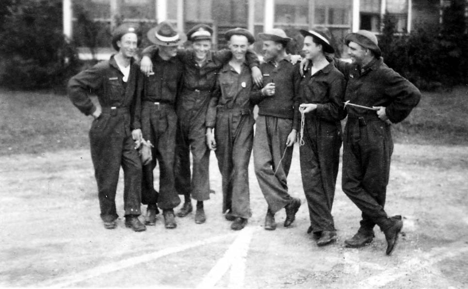A group of seven smiling flight cadets in dark coveralls and campaign hats standing casually together with their arms around each other's shoulders.