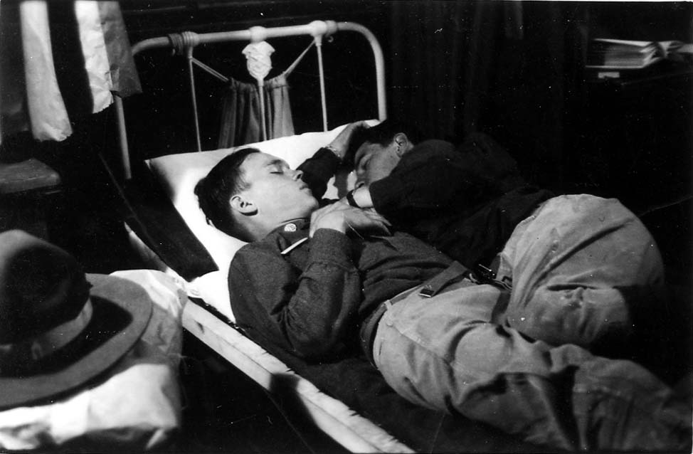 Two fully uniformed flight cadets sleeping closely together on a single narrow cot in the barracks, demonstrating profound exhaustion.
