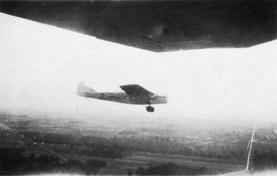 An air-to-air photograph taken from beneath the wing of another aircraft, showing a Ford Tri-Motor passenger plane flying over a rural landscape.