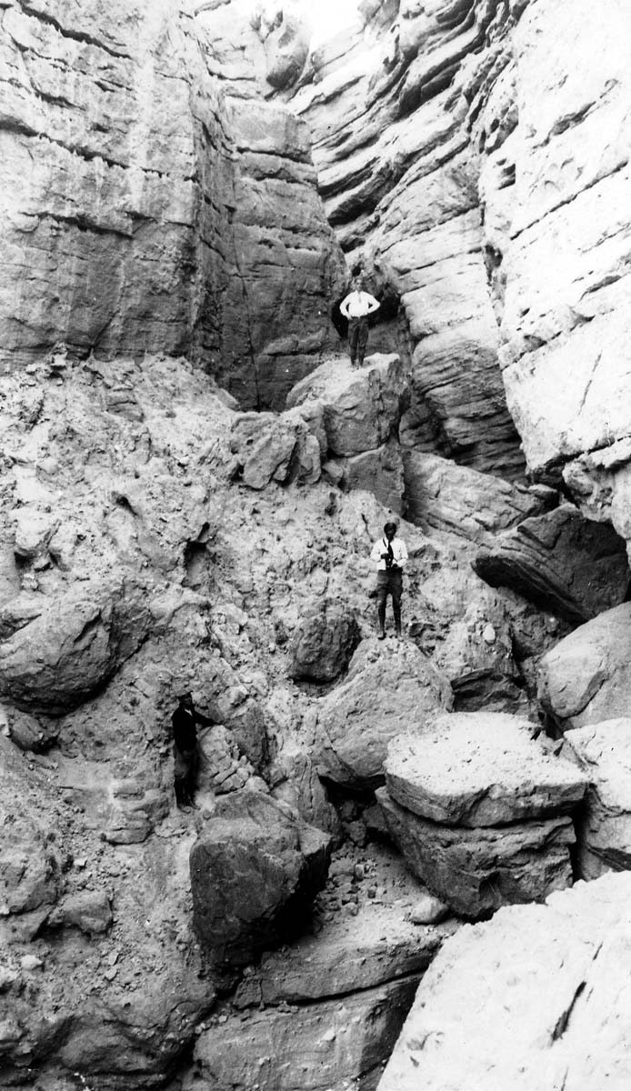 Three men, one holding a camera, exploring a deep, rugged canyon filled with massive boulders and steep rock walls.