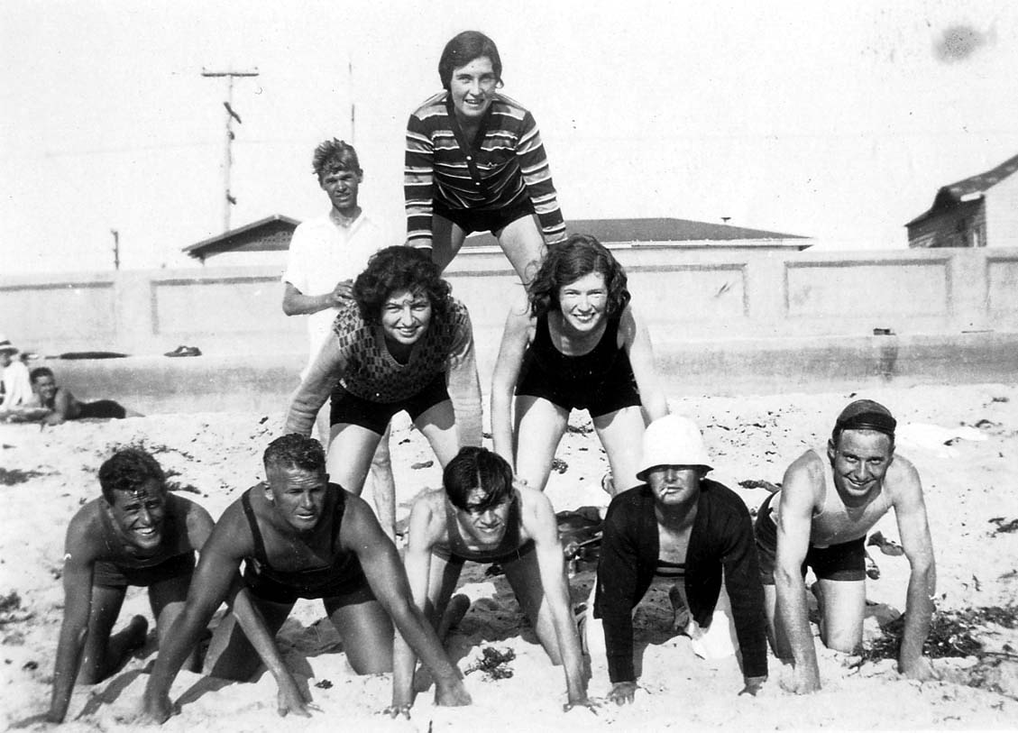 A group of eight friends in 1920s swimwear forming a playful human pyramid on the sandy beach, with a seawall in the background.