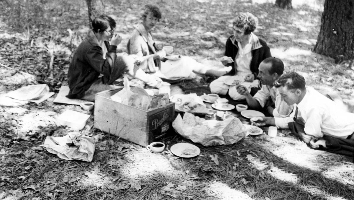 A group of five friends enjoying a picnic in a sunny, wooded area, sitting on a blanket around a wooden box filled with food.