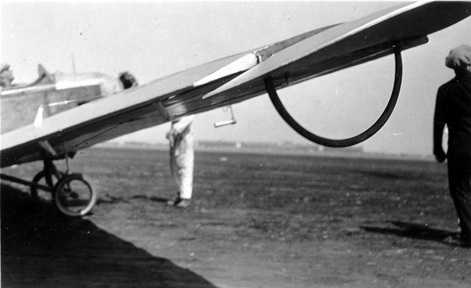 A close-up, low-angle view showing the underside of an aircraft wing, focusing on a curved metal handling bar and the structural ribs beneath the fabric.