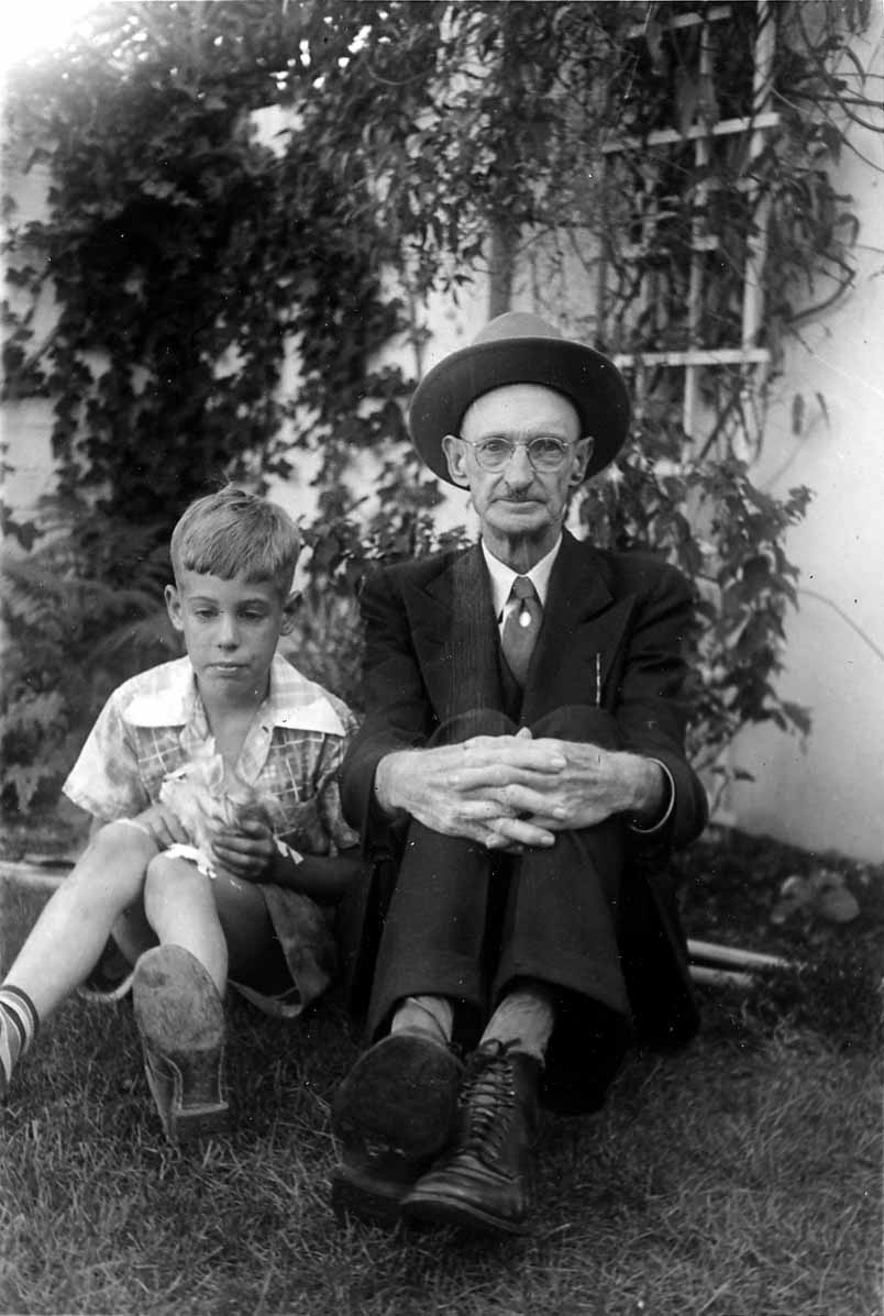 A young boy sitting in the grass next to an older gentleman wearing a dark suit, tie, glasses, and a fedora hat.