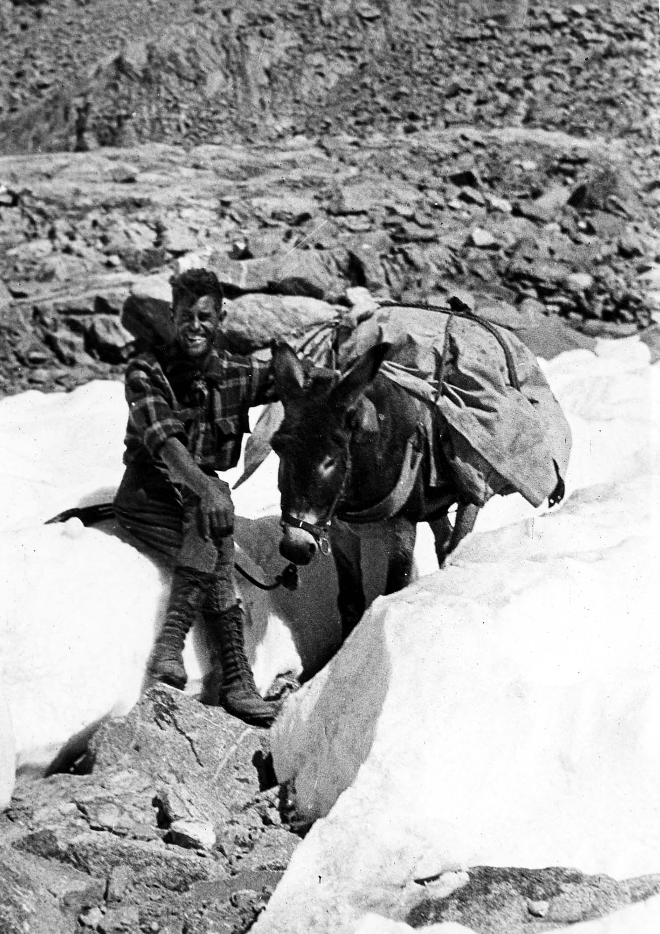 A smiling Donald A. Hall Sr. sitting on a patch of high-altitude snow next to a loaded pack mule.