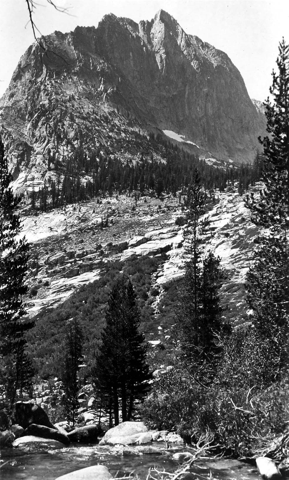 A sheer, dramatic granite peak towering vertically over a boulder-strewn alpine creek and dense pine forest in the Sierra Nevada.