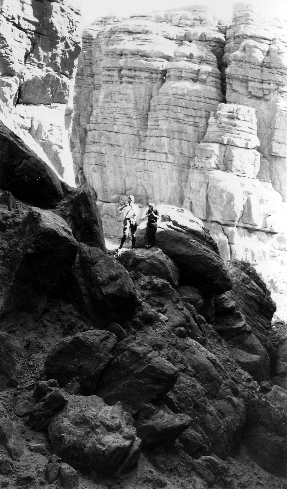 Two hikers standing atop massive, dark boulders in a deep, rugged canyon gorge with towering rock walls behind them.