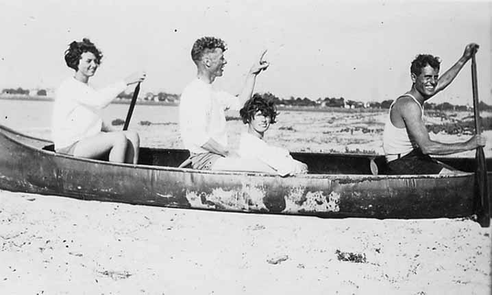 Two men and two women sitting inside a canoe that is resting entirely on dry beach sand, holding paddles.