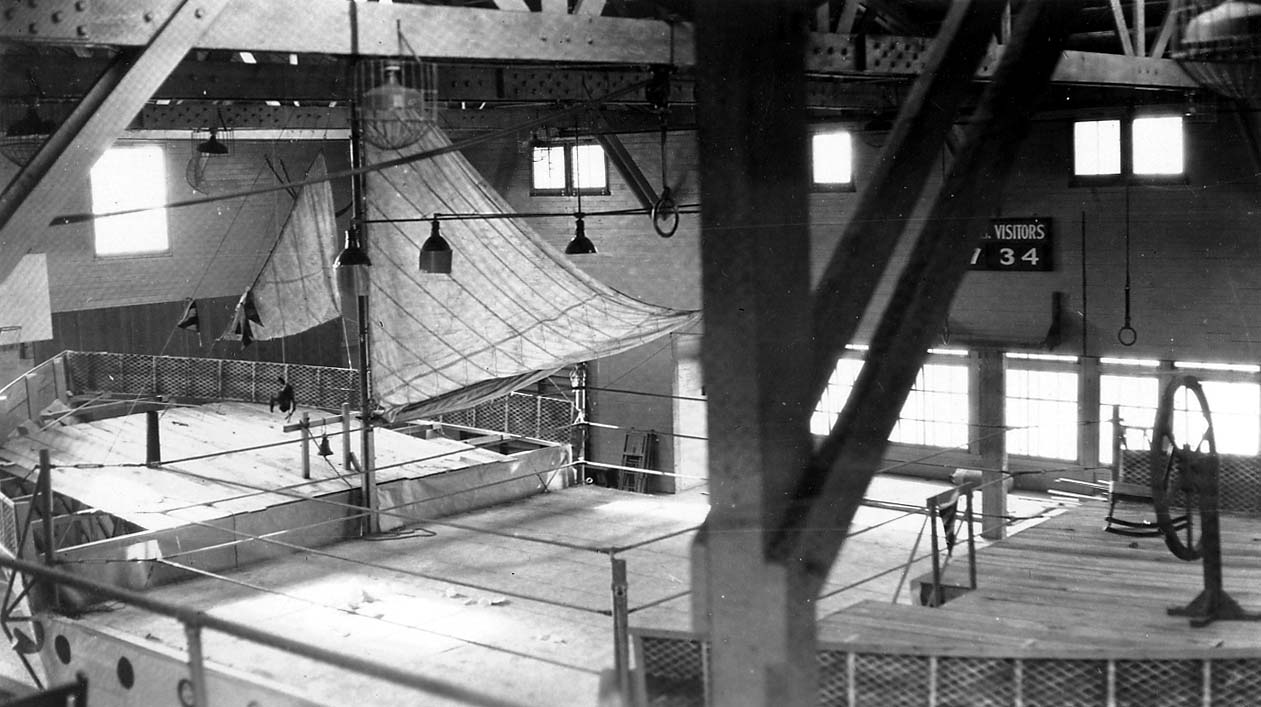A massive timber-trussed indoor gymnasium featuring an elevated boxing ring beneath a suspended canvas awning and a wall scoreboard.