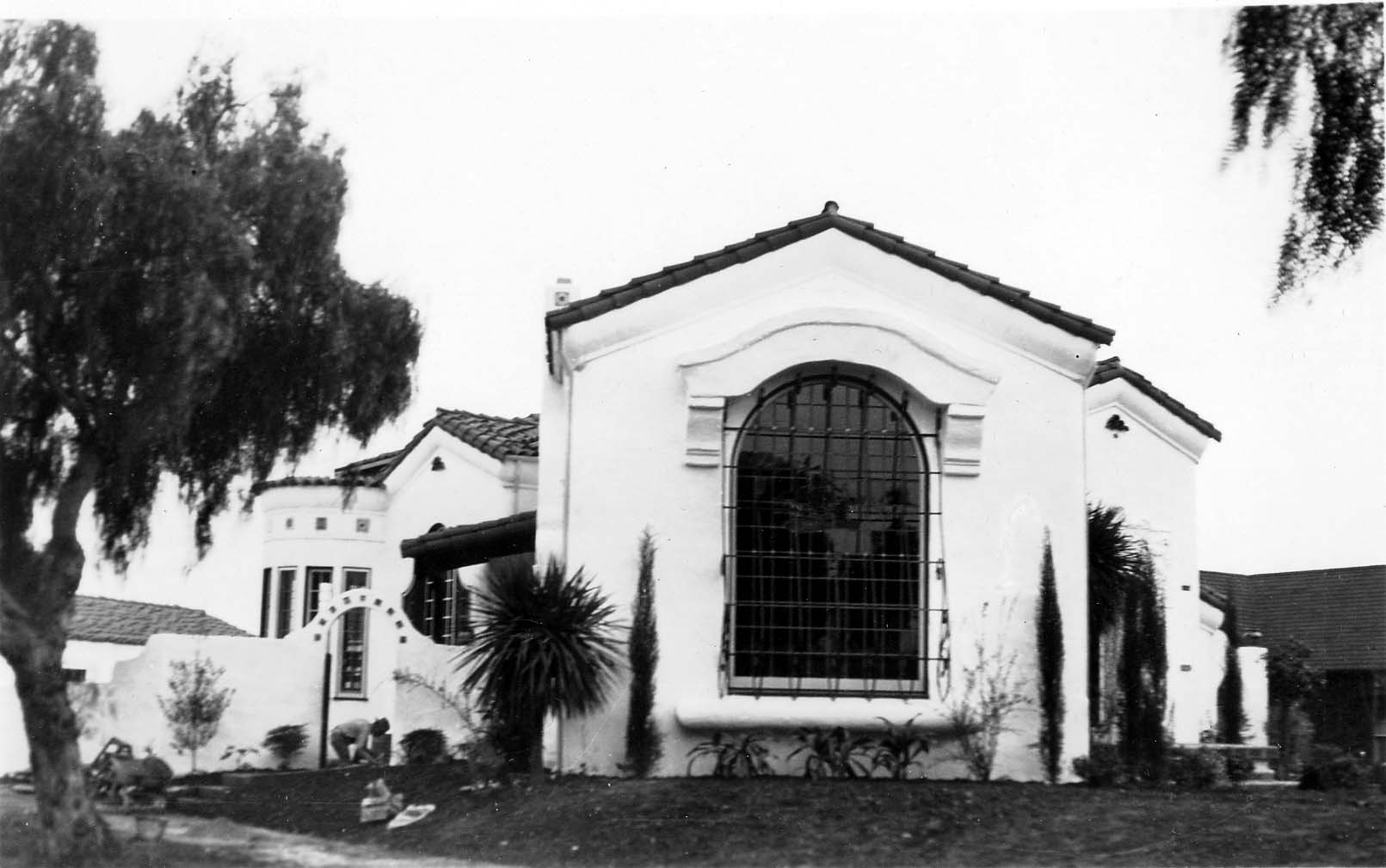 An exterior view focusing on a massive, arched, iron-grated window set into the white stucco wall of a wealthy Spanish Colonial Revival home.