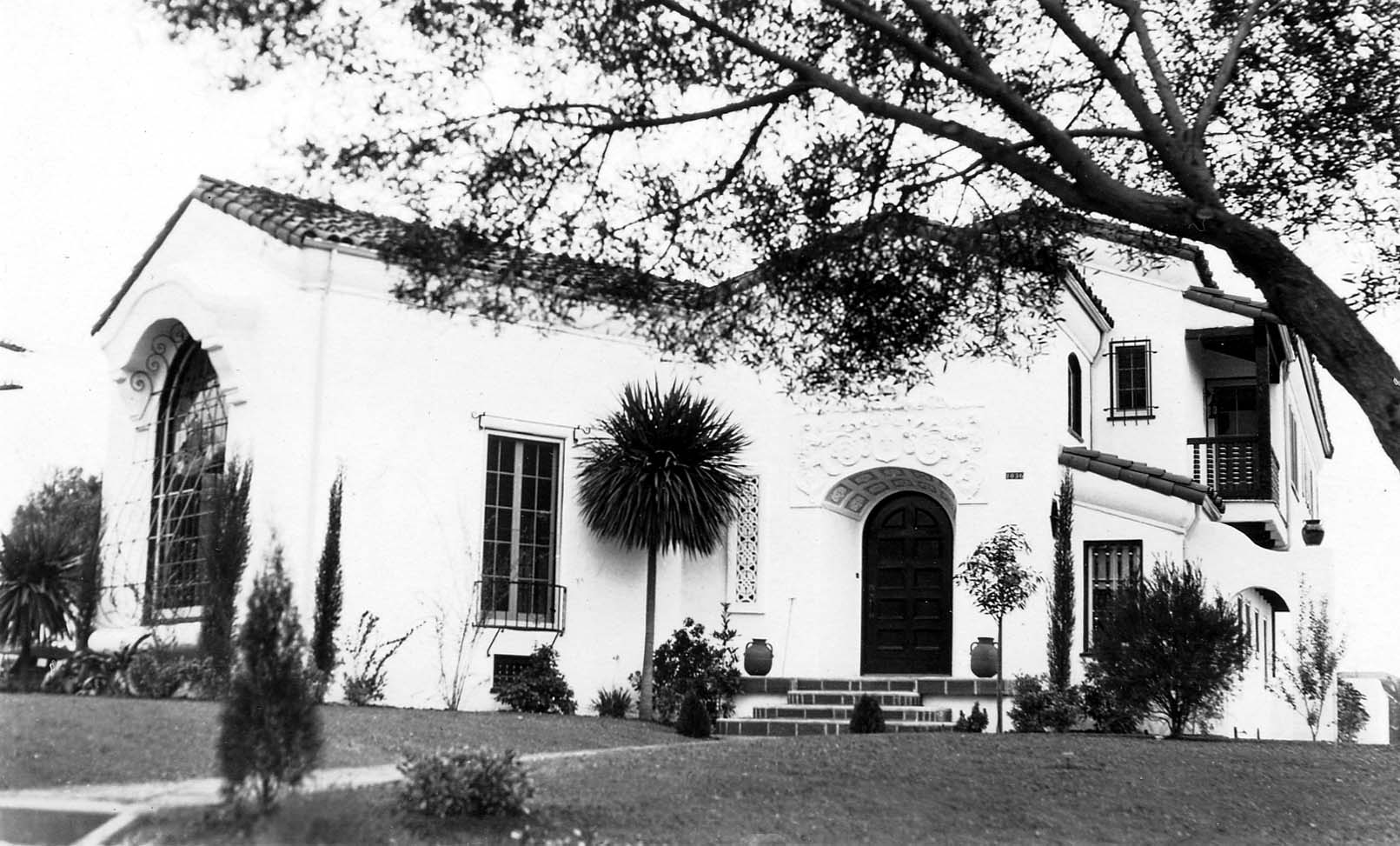 A wide exterior shot of a large, wealthy estate featuring Spanish Colonial Revival architecture, a manicured lawn, and a prominent arched entryway.