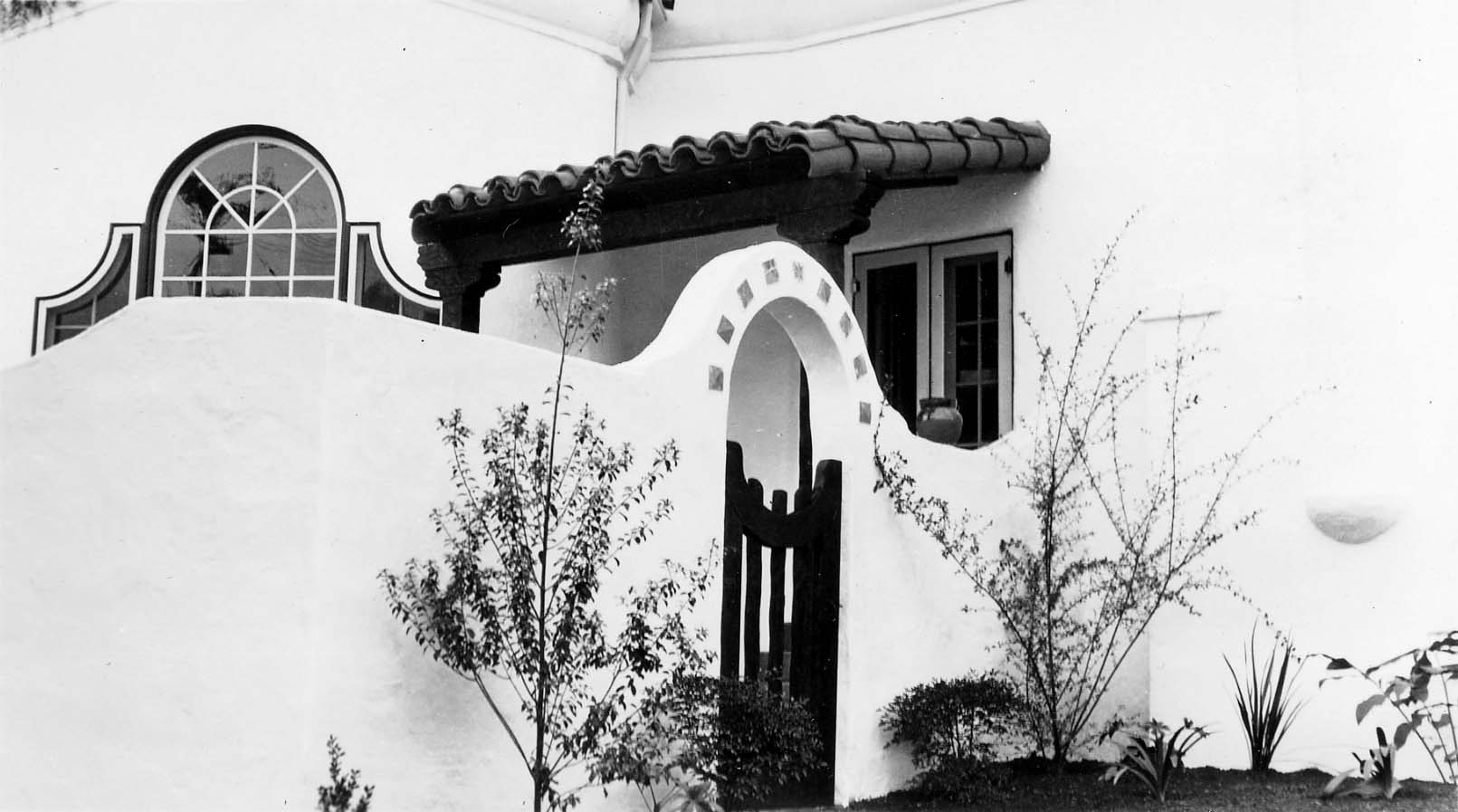 An exterior architectural view looking through a white stucco archway toward a dark wooden door and a tiled patio with potted plants.
