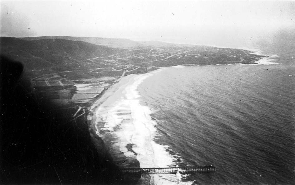 An aerial perspective looking south along the San Diego coastline, showing the Scripps Institution of Oceanography Pier, the crescent of La Jolla Shores, and La Jolla Cove.