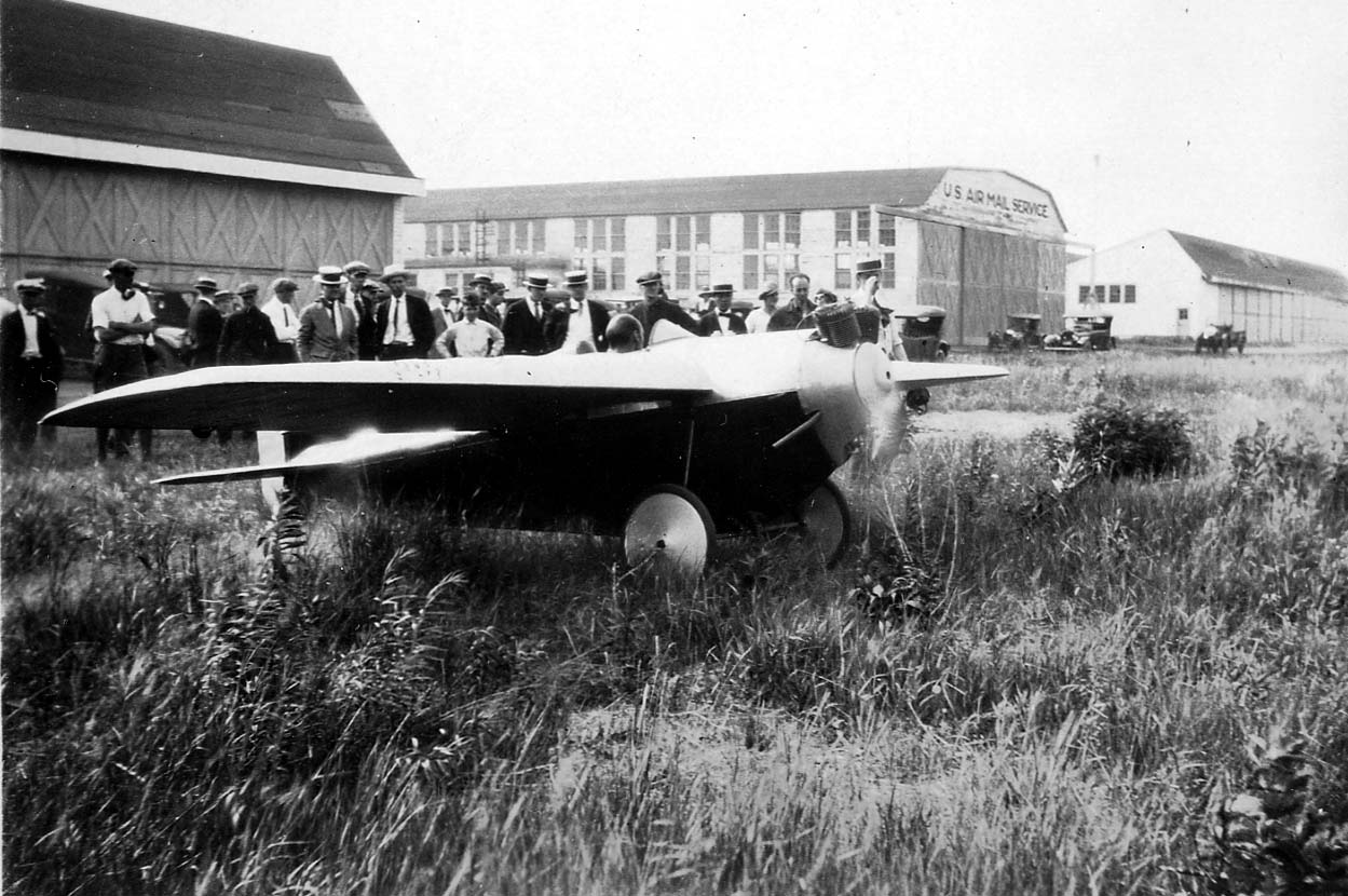 A sleek single-seat monoplane racer with a two-cylinder engine parked in weeds outside a US Air Mail Service hangar before a crowd.