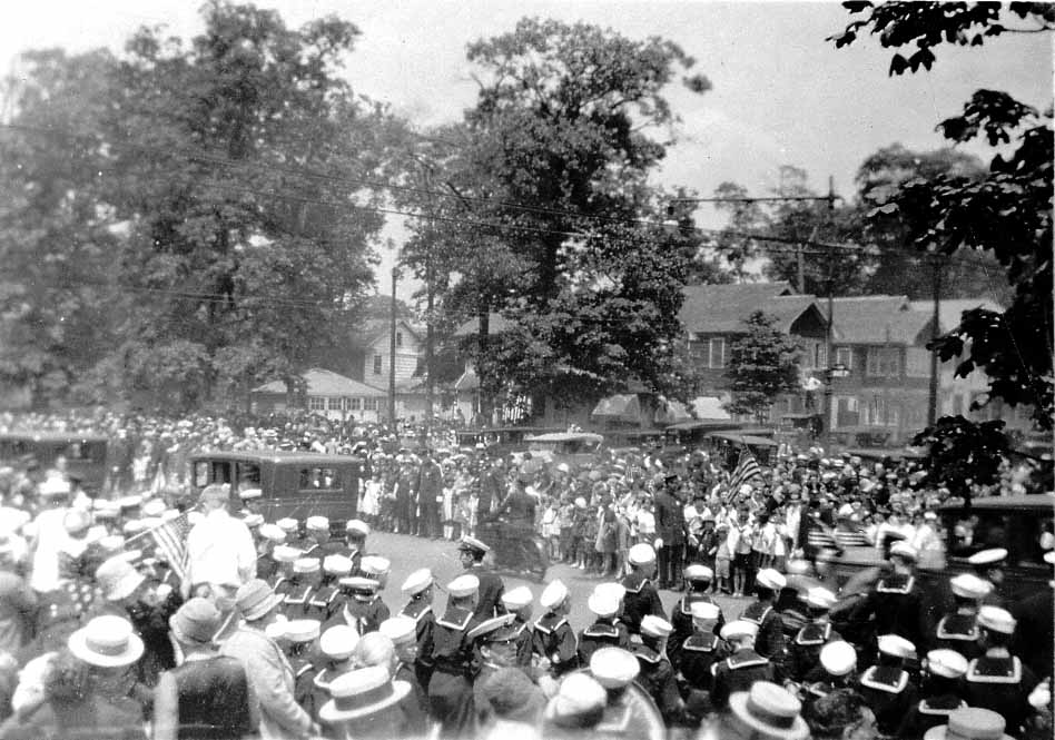 A wide view of a massive celebration parade, featuring a large crowd, automobiles, and Navy sailors lined along a residential street.