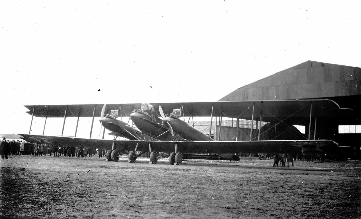 A massive crowd gathered around the three-engine LWF Model H Owl biplane bomber parked in front of a giant airfield hangar.