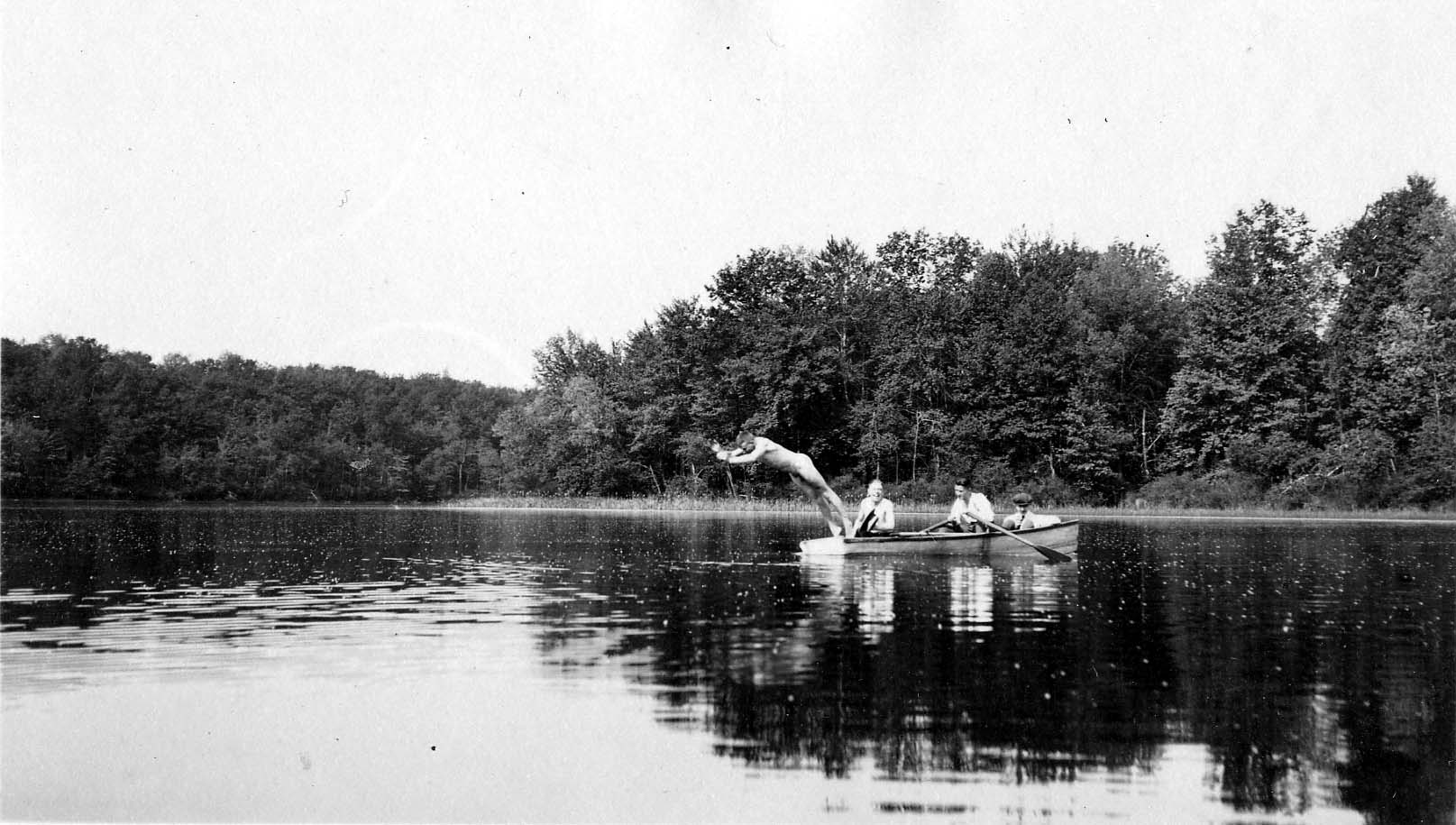 A man diving headfirst from a wooden rowboat into a calm, tree-lined lake, while two other men sit in the boat holding the oars.
