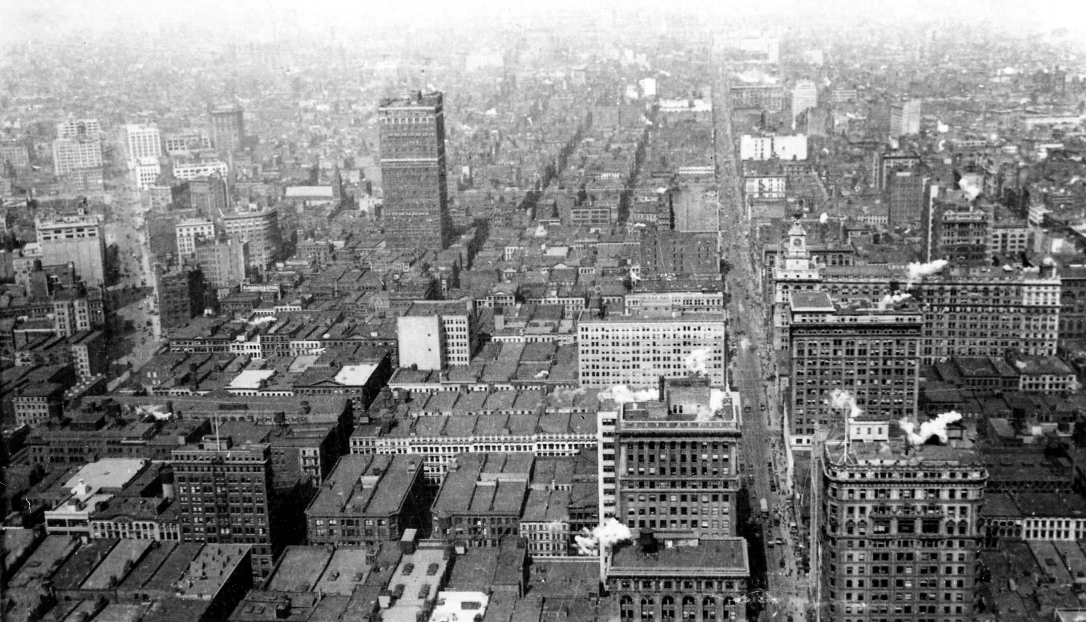 An expansive elevated panorama looking north across Manhattan's commercial avenues, rooftop water tanks, and steam vents from the heights of the Woolworth Building.