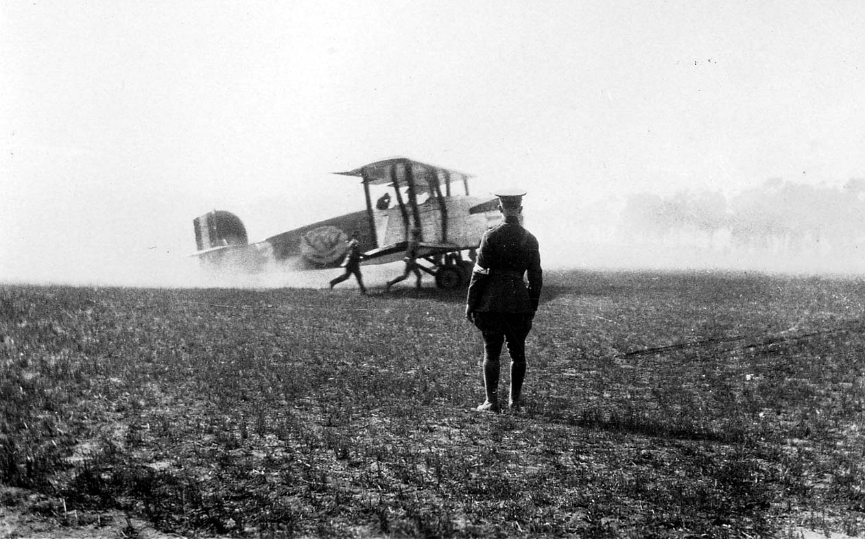 A military biplane kicking up dust on a tarmac as crew members rush toward it, watched by a military officer in uniform.