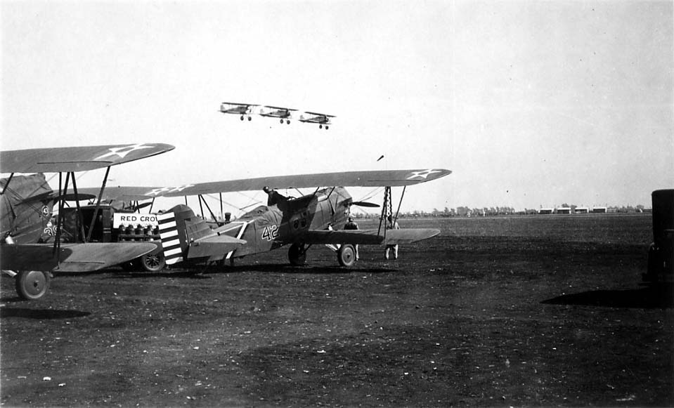 A low-angle view of three military biplanes flying in formation over several parked aircraft on a dirt airfield.