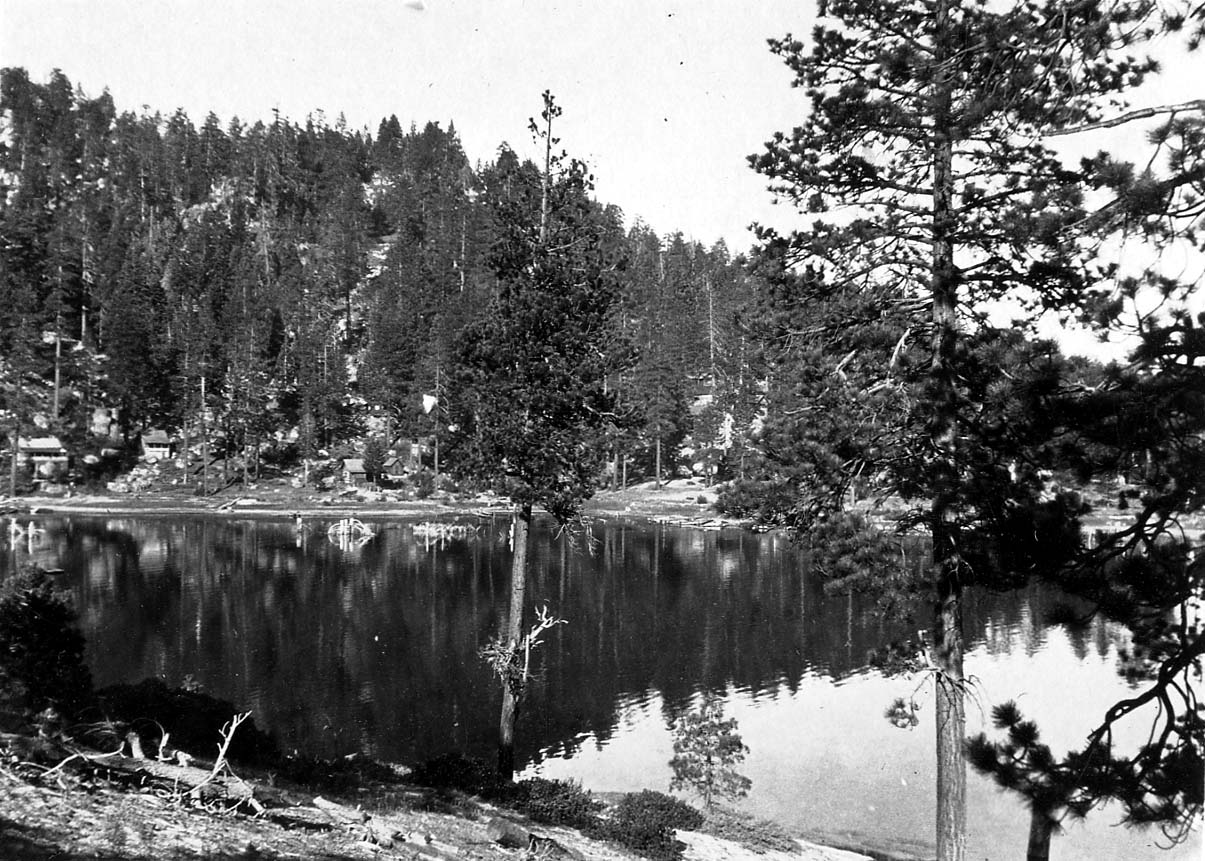A glassy, mirror-like mountain lake reflecting a dense pine forest and scattered small cabins along the hillside.