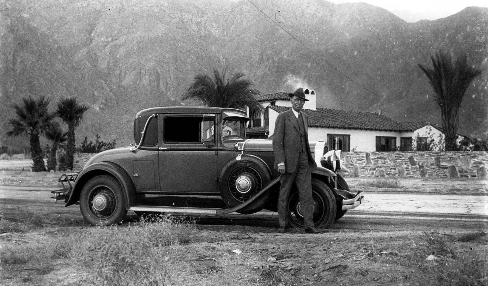 A distinguished man in a three-piece suit standing beside a luxury coupe in front of a Spanish Colonial home with desert mountains in the background.