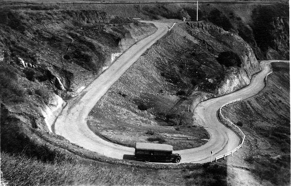 A vintage passenger motorcoach navigating a sharp, unpaved hairpin switchback descending the Santa Monica coastal bluffs.