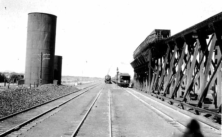 A steam train in the distance passing between large cylindrical water towers and a massive wooden industrial loading trestle.