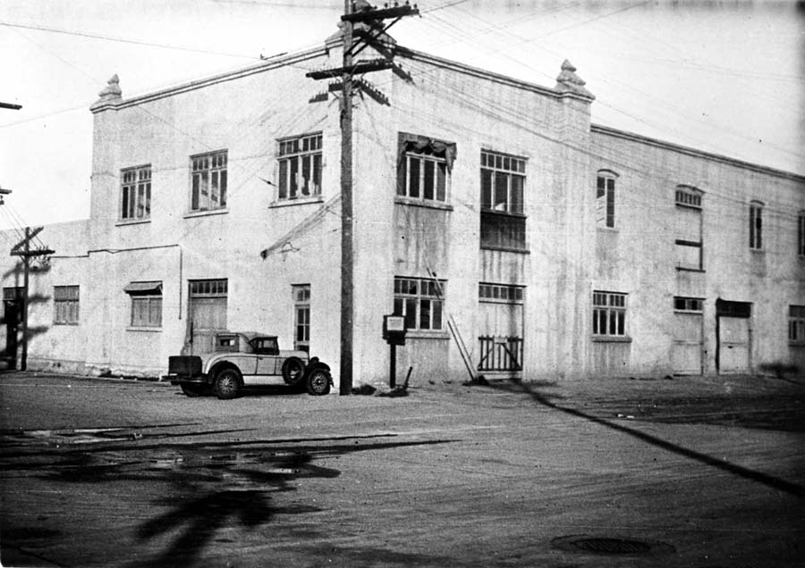 A large, multi-story, whitewashed industrial building on a corner lot, originally a fish cannery and later the Ryan Airlines factory, with a vintage vehicle parked outside.