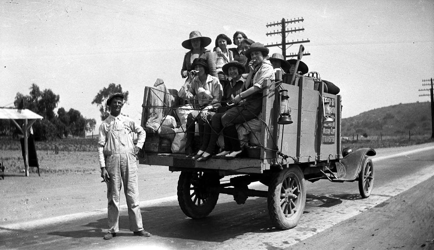 A vintage Mission Cliff Transfer flatbed truck carrying a group of young men and women in 1920s attire and camping gear, with a driver in overalls standing beside the vehicle on a paved road.