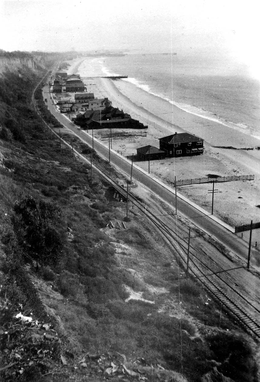 A high-angle elevated panorama of the Santa Monica shoreline showing railroad tracks, the coast road, and rows of dark wooden beach cottages on the sand.