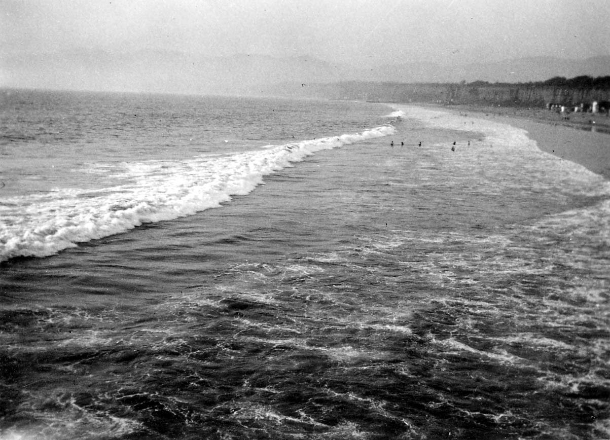 A high-angle view looking over rolling ocean waves with swimmers in the surf along the wide sandy beach of Santa Monica, with coastal palisades in the background.