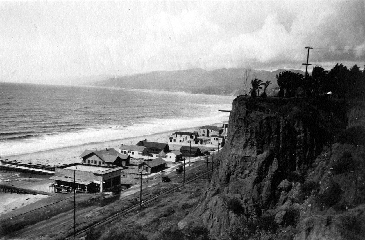 A steep vertical perspective looking down a sheer Palisades cliff face to coastal railroad tracks, beach bungalows, and the Pacific Ocean.