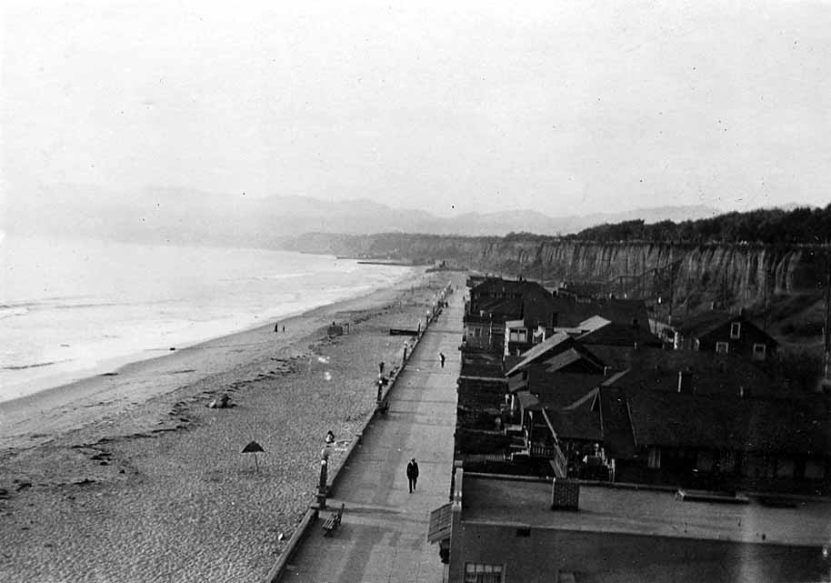 A high-angle perspective looking down the long paved pedestrian Ocean Front Walk lined with Craftsman beach bungalows, vintage streetlamps, and a lone walker.
