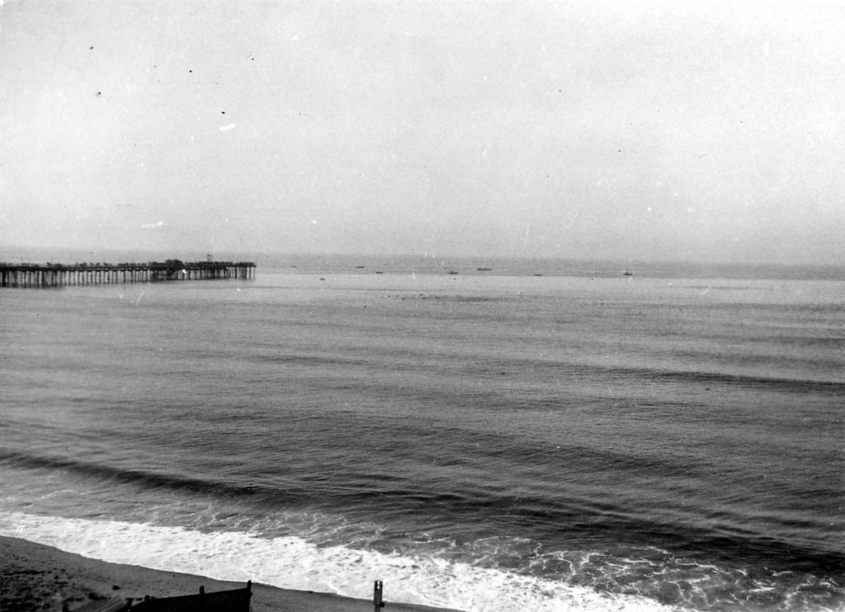 A high-angle perspective looking down at long, rhythmic ocean swells rolling toward the beach, with a long wooden pleasure pier extending into the sea on the left.